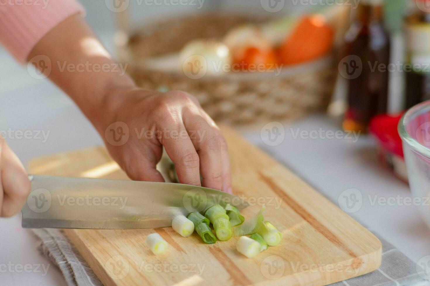 Female hand using a knife to cut Negi Japanese Long Onion or Japanese Scallion or Green Onion on wooden cutting board. Woman preparing food in the kitchen at home. photo