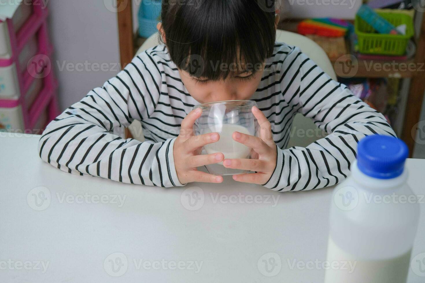 Cute Asian girl drinking a glass of milk at home in living room. Little girl drinking milk in the morning before going to school. Healthy food in childhood. photo