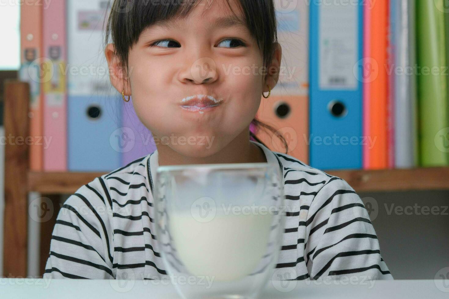 Cute Asian girl drinking a glass of milk at home in living room. Little girl drinking milk in the morning before going to school. Healthy food in childhood. photo