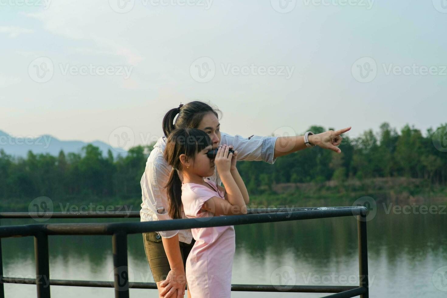 Mother teaching her little daughter to use binoculars by a reservoir in the summer garden. Explore and adventure concept. photo