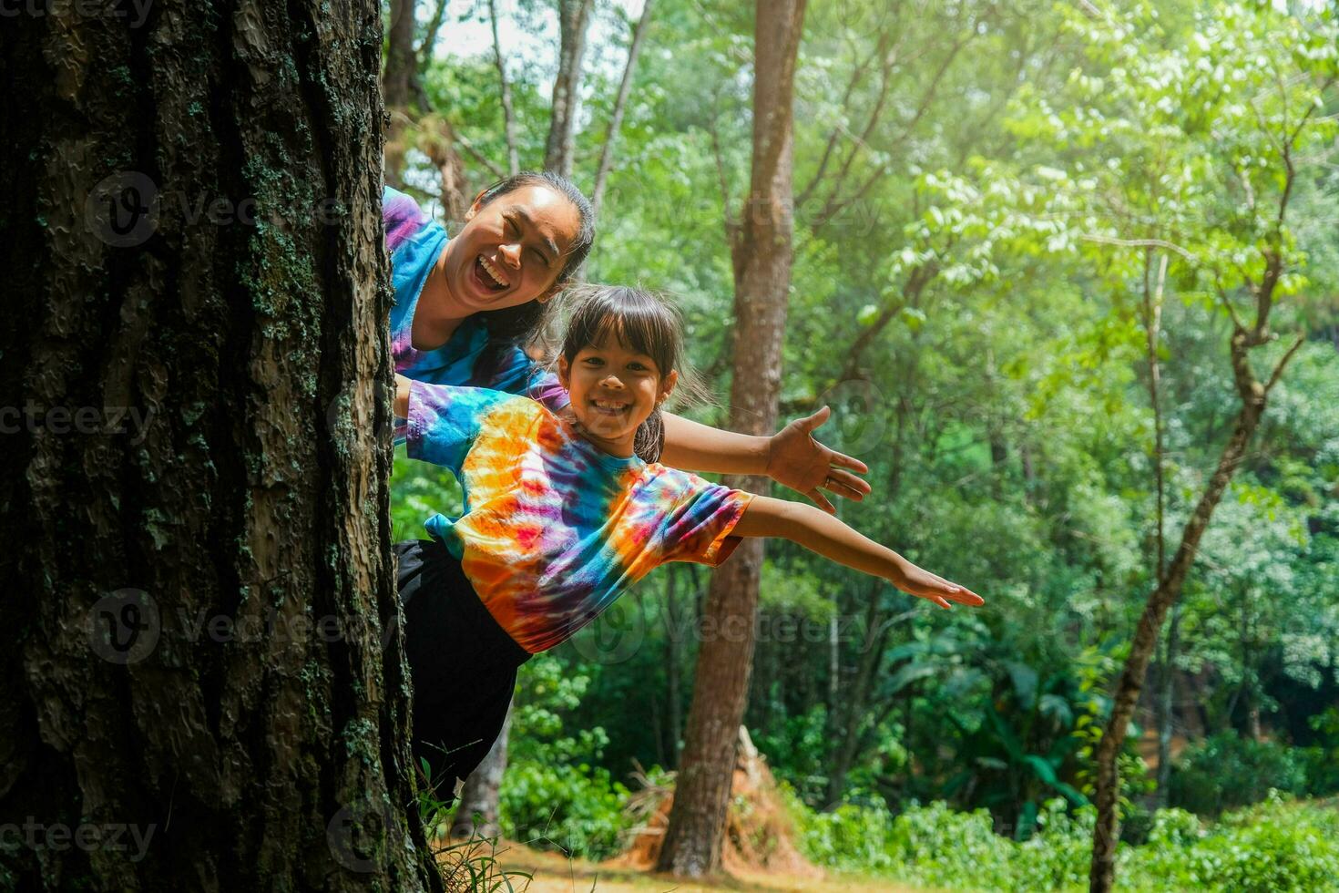 Happy family hiding behind a tree while playing in the park. Happy mother and daughter hiding behind a tree trunk. Green environmentally friendly lifestyle. Love and protect nature concept. photo