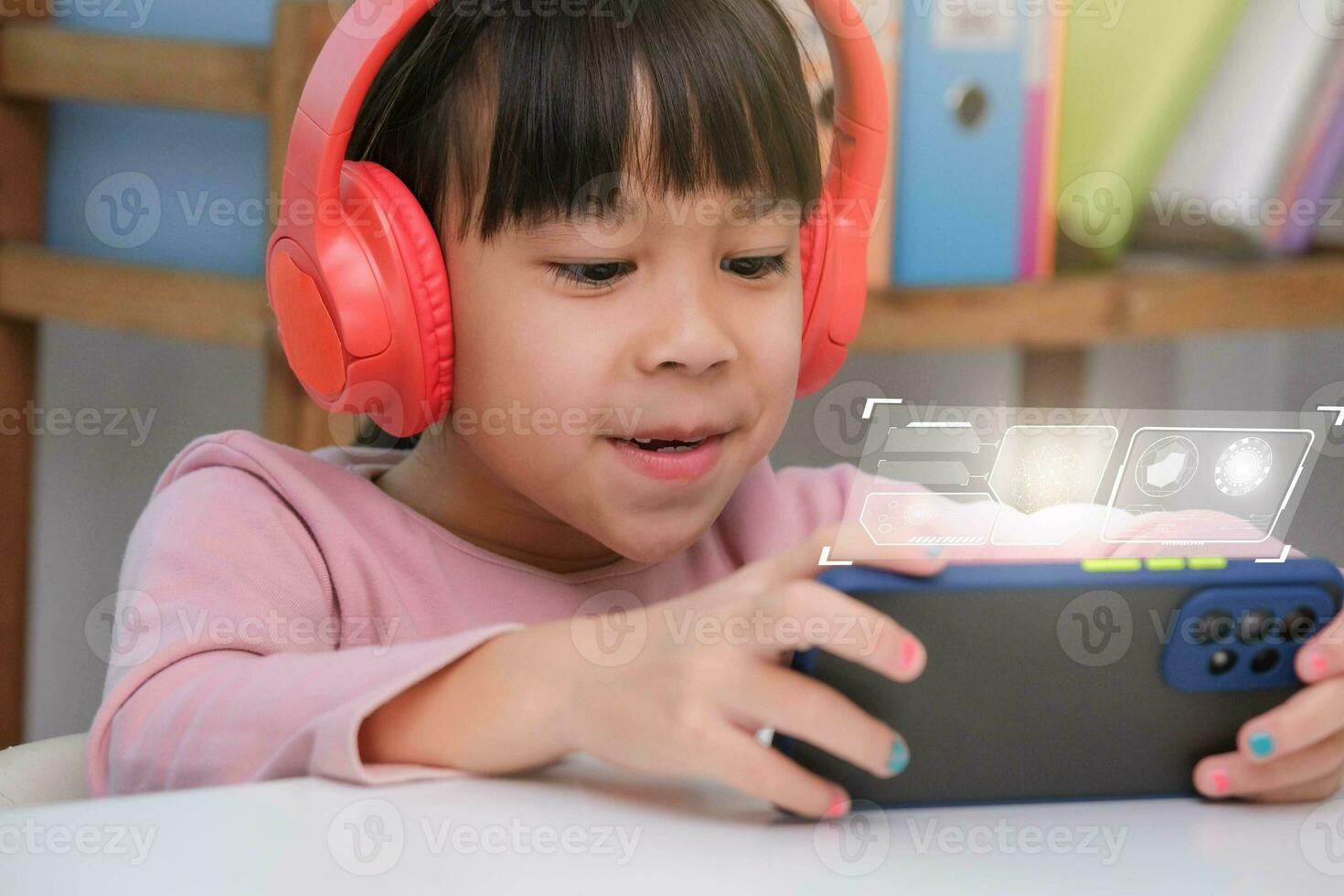 Cute elementary school girl wearing headphones holding a smartphone. Happy Asian kid studying online on smartphone or homeschooling, listening to music or playing games. photo