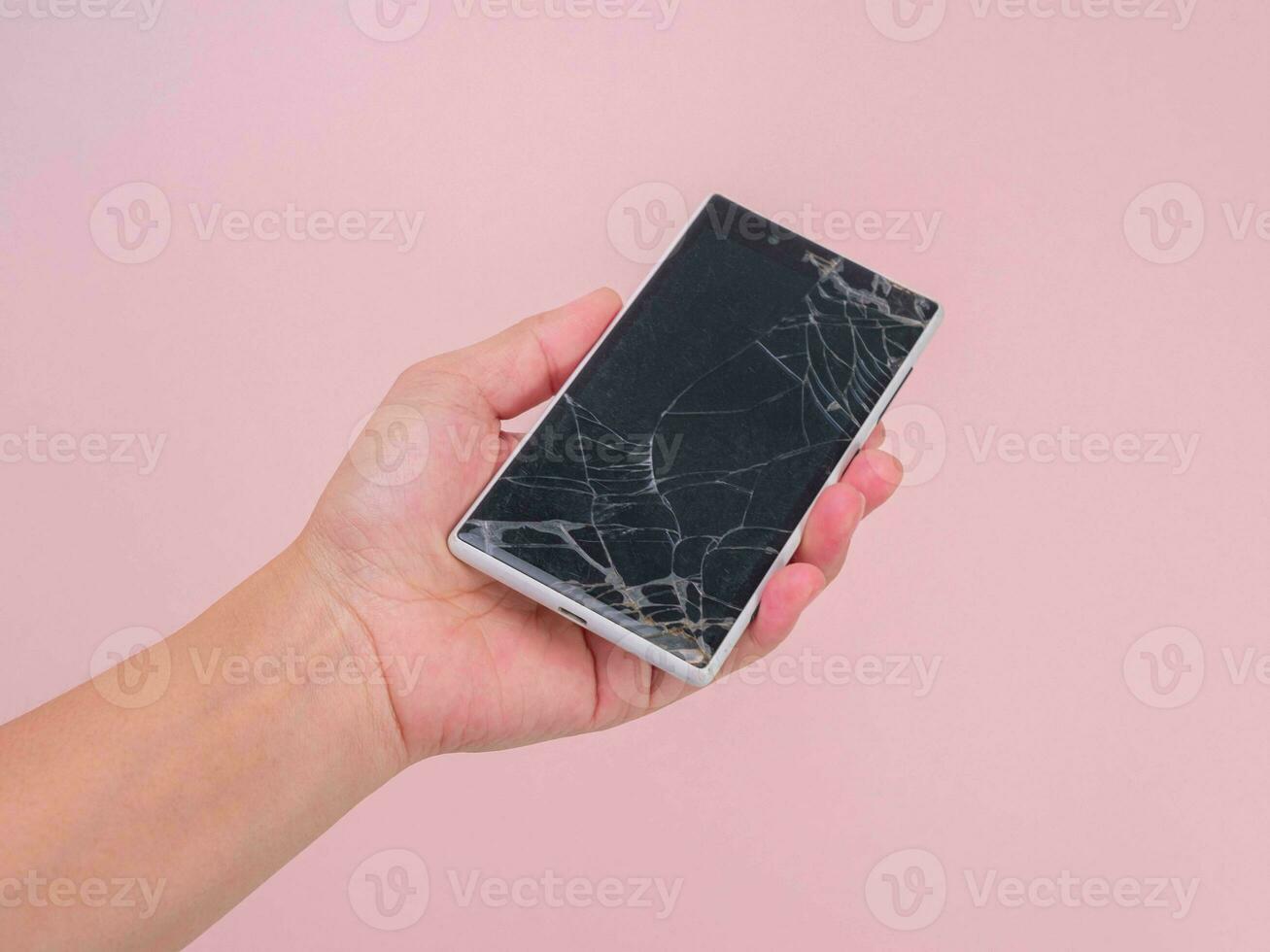 Close-up of hand holding mobile phone with broken touch screen on pink background. Female hand holding old phone with cracked and damaged screen. photo