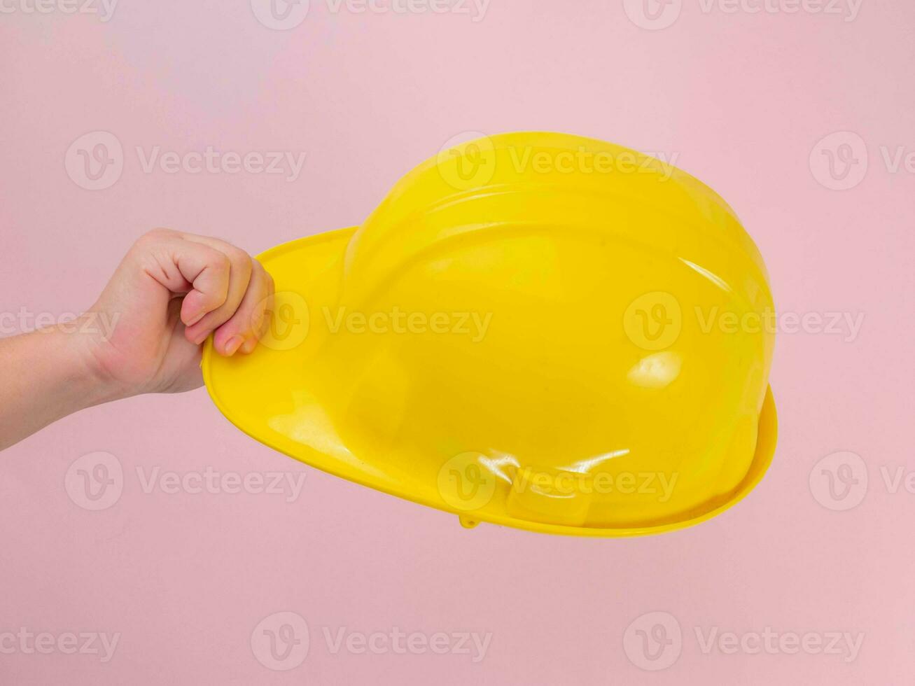 Close-up of female engineer hand holding yellow safety helmet on pink background. Human hand holding a hard hat. photo