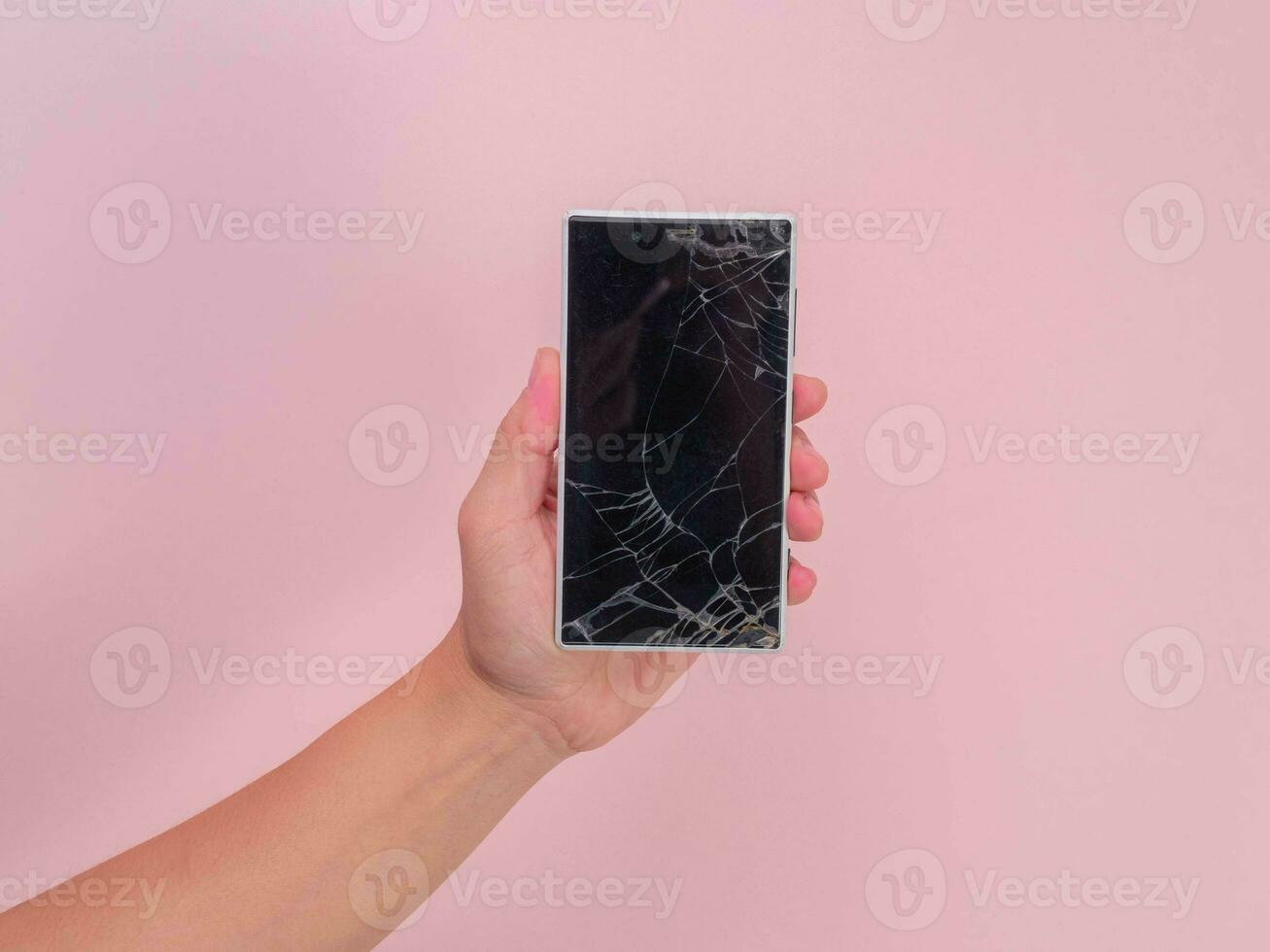 Close-up of hand holding mobile phone with broken touch screen on pink background. Female hand holding old phone with cracked and damaged screen. photo