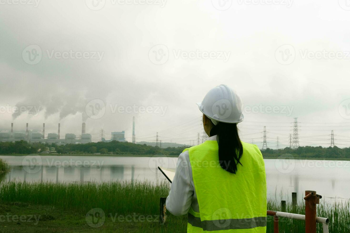 hembra jefe ingeniero vistiendo un verde chaleco y casco soportes fuera de en contra el antecedentes de carbón poder planta estación y vapor en el Mañana neblina. ingeniero trabajando y ordenador portátil a carbón poder planta. foto