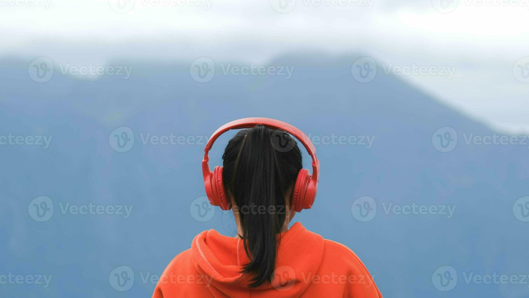 vista trasera de una mujer joven parada en una colina tranquila y escuchando música con auriculares por la mañana. mujer con un suéter disfrutando de la belleza de la naturaleza mirando la montaña en invierno. foto