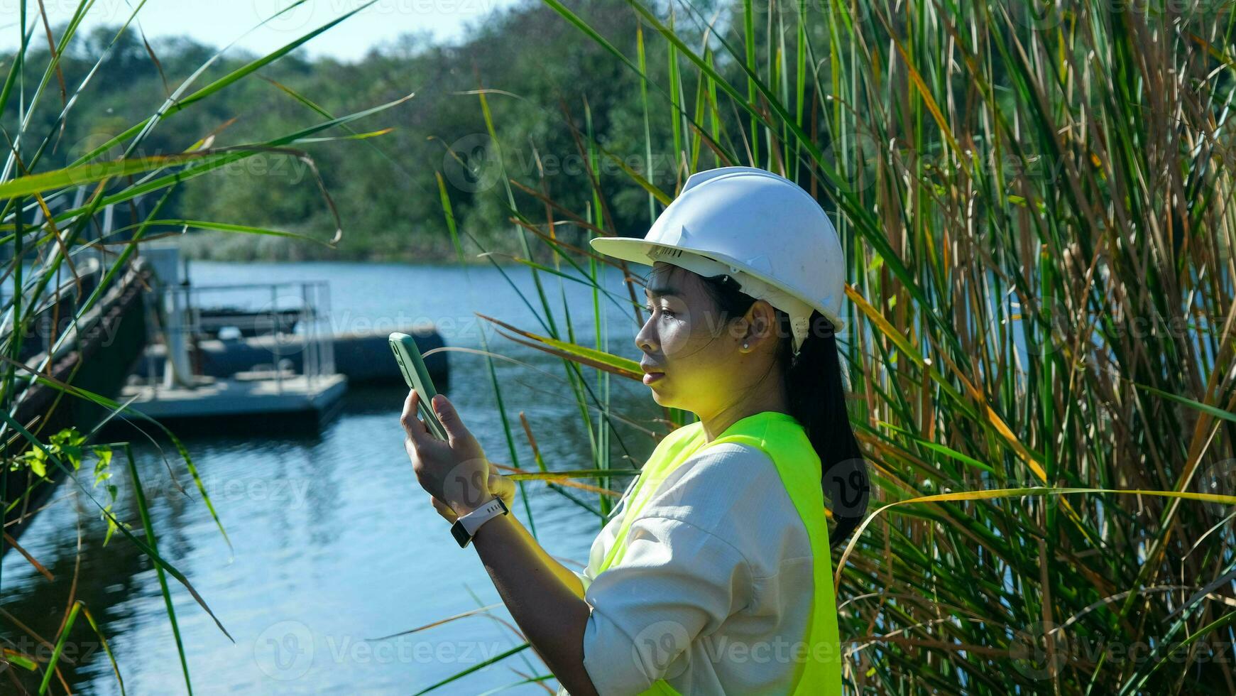 Environmental engineer wearing a white helmet uses a mobile phone to record data analyzing oxygen levels in a reservoir. Water and ecology concept. photo