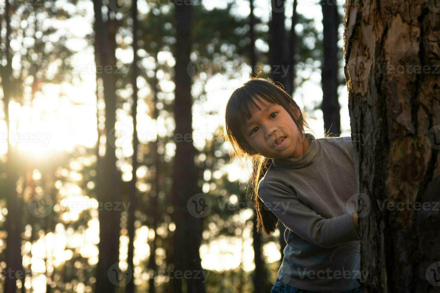 Funny little girl peeking out from behind tree in winter park. Cute smiling child playing hide and seek in the forest. Children look into the camera and smile. Happy family on vacation in nature. photo
