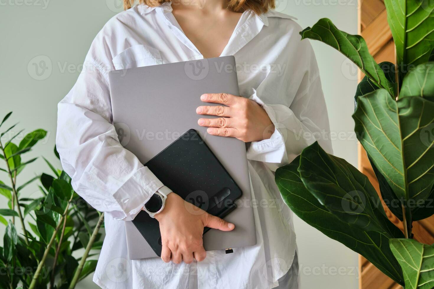 A woman holds a laptop and graphics tablet against a background of greenery. photo