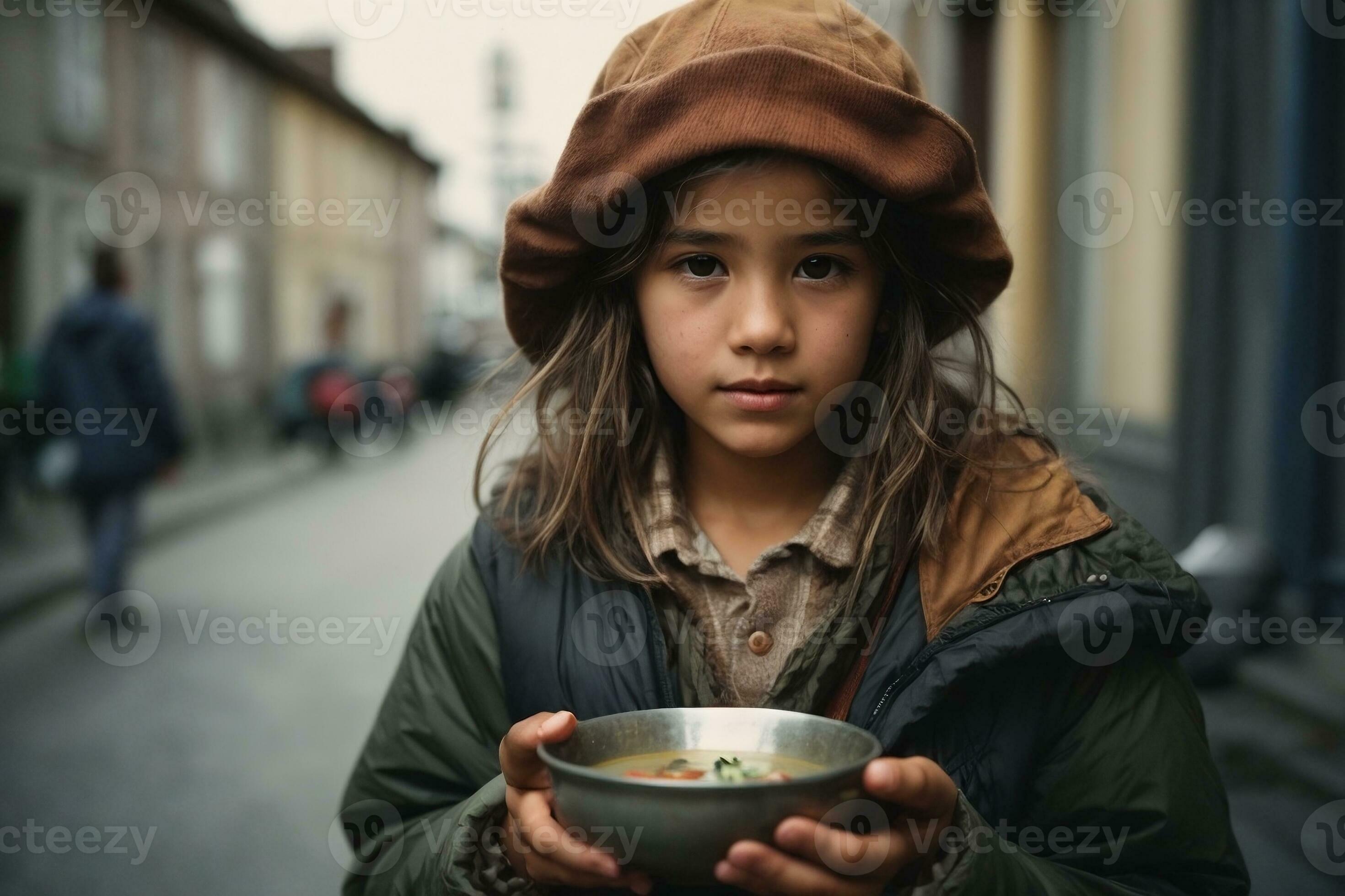 Photo of a homeless girl in old clothes holds a metal bowl of