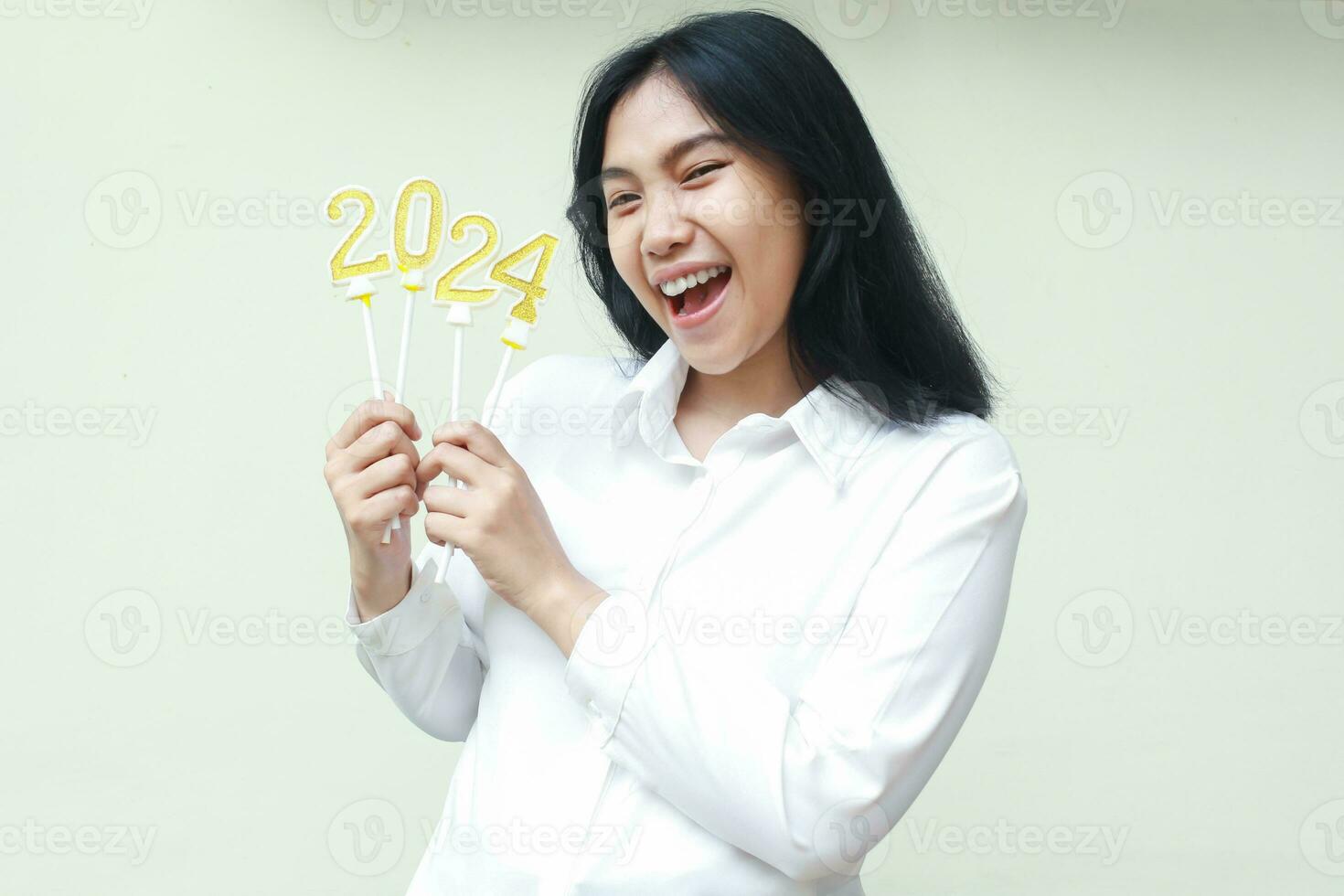retrato de emocionado asiático joven mujer riendo a cámara celebrando 2024 nuevo años víspera por participación número dorado velas vistiendo blanco formal camisa aislado por blanco foto