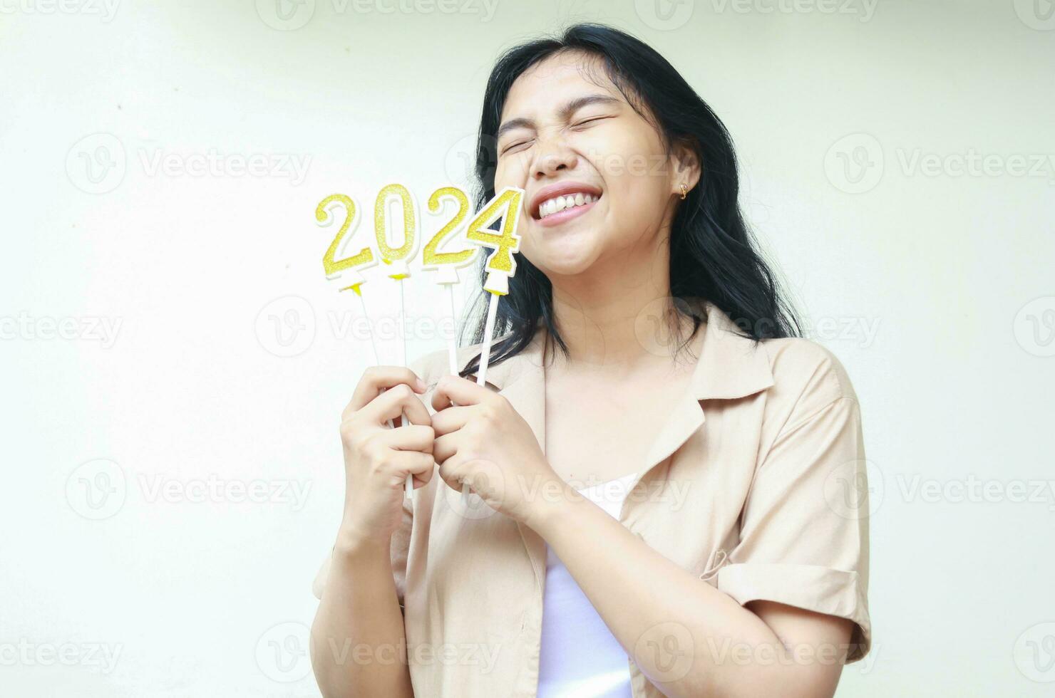 asian young woman excited to celebrating new years eve with holding golden candles numbers 2024 and closed eye wearing casual brown shirt isolated by white background photo