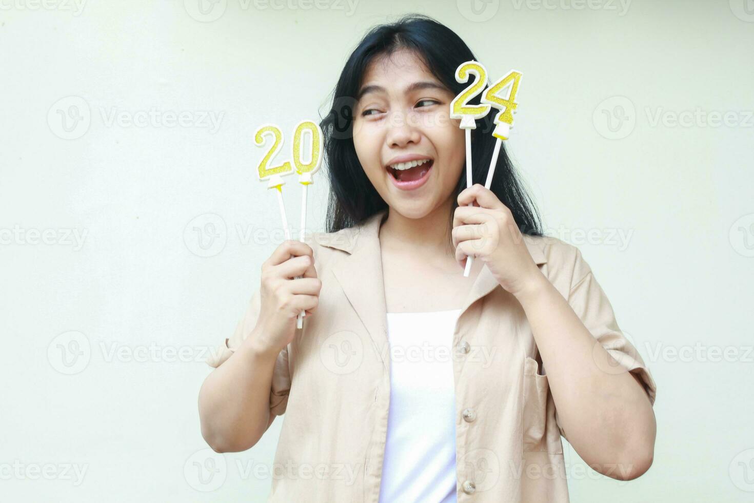 portrait of excited young woman asian holding golden candles numbers 2024 with white stick to celebrating new years eve wearing casual brown shirt looking away isolated on white photo
