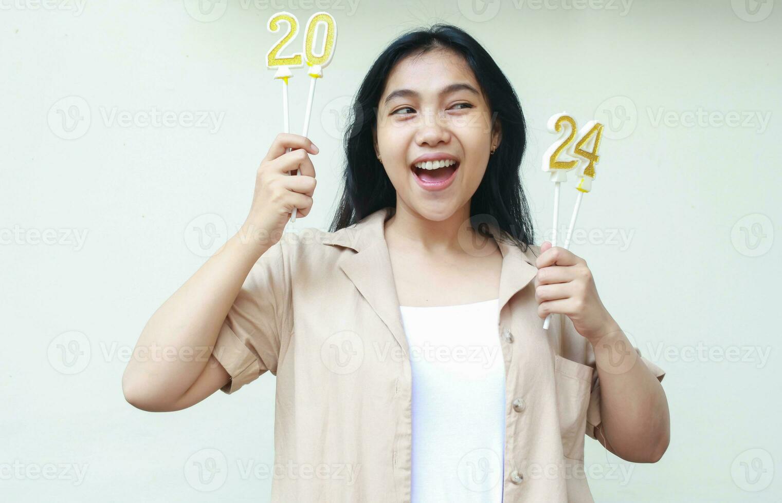 cheerful asian young woman smiling and holding gold candles numbers 2024 looking away wear casual brown shirt isolated in white. new years eve concept photo