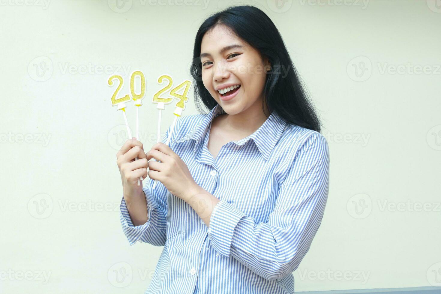 excited successful asian young business woman holding golden candles numbers 2024 for new years eve celebration wearing stripes blue shirt looking to camera isolated on white background photo