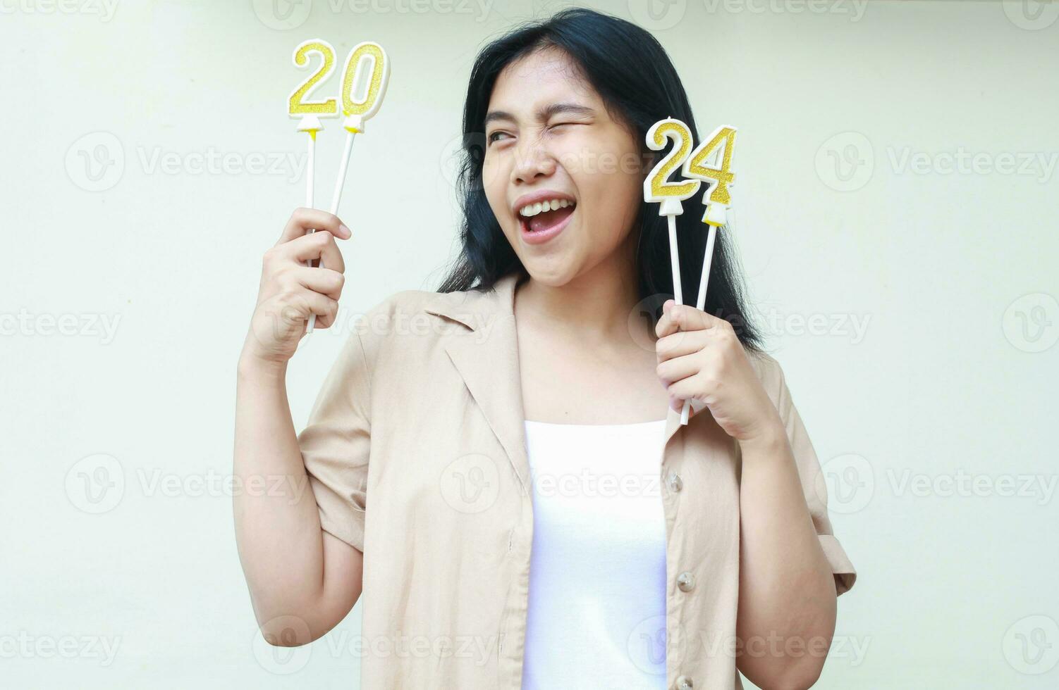 excited asian young woman holding gold and white candles number 2024 looking side wearing casual shirt isolated by white background photo