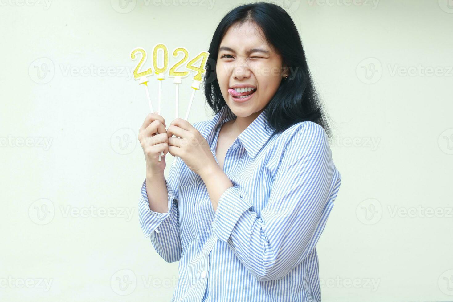 emocionado asiático joven negocio mujer sonriente con uno ojo parpadeo participación dorado velas números 2024 a celebrando nuevo años víspera vestir azul rayas camisa mirando a cámara aislado en blanco antecedentes foto