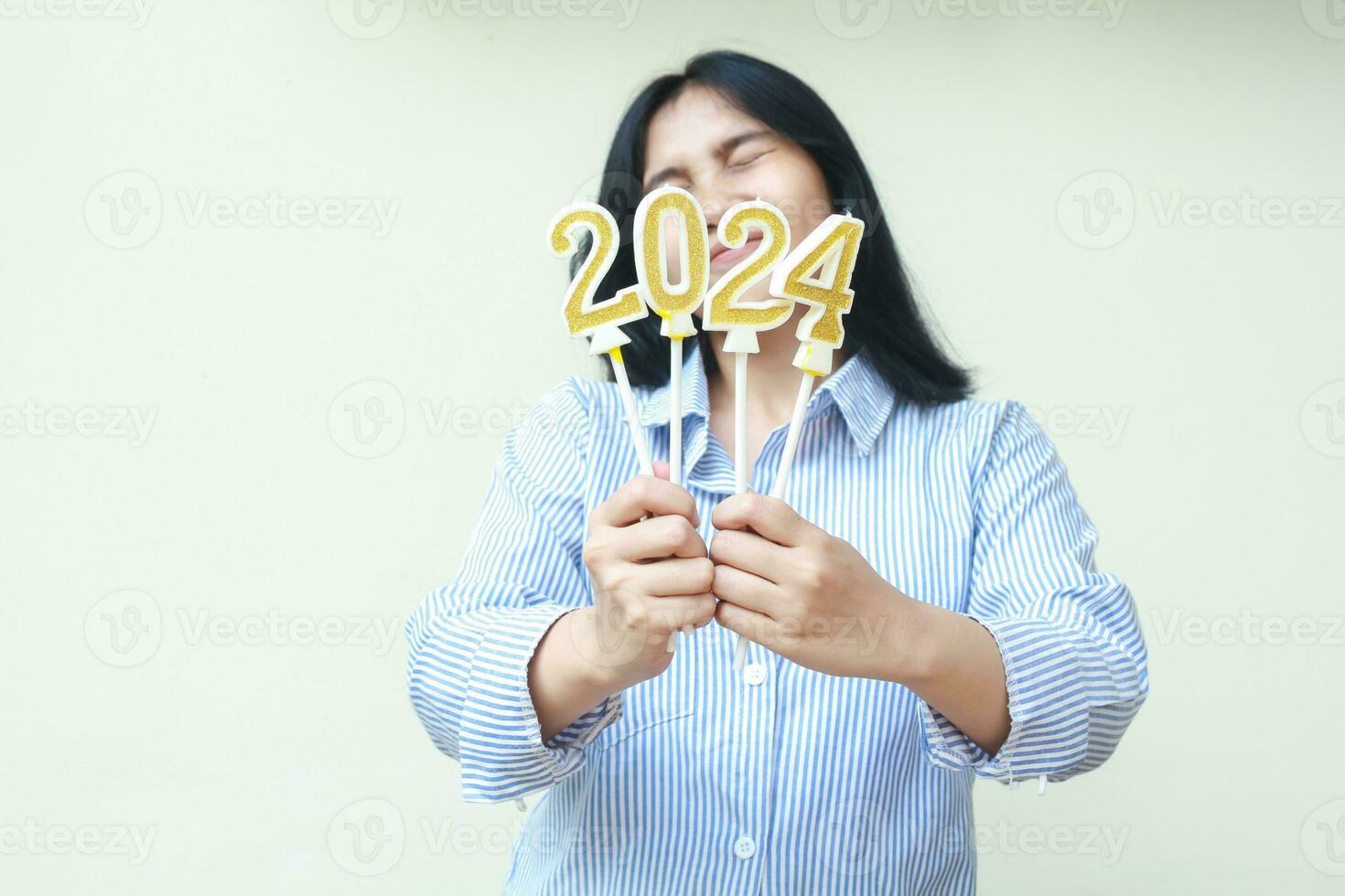 retrato de contento asiático joven negocio mujer cerca su ojos y cubierta cara con dorado velas número 2024 a celebrar nuevo años víspera vistiendo azul rayas camisa aislado foto