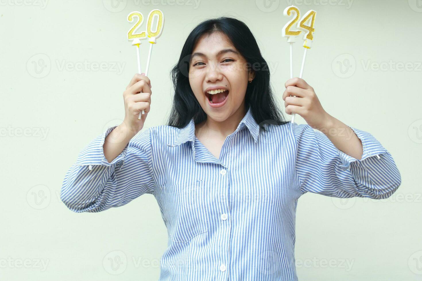 retrato de emocionado asiático joven mujer gritos a anunciar nuevo años víspera con sostener dorado velas números 2024 vistiendo azul rayas camisa mirando a cámara aislado en blanco antecedentes foto