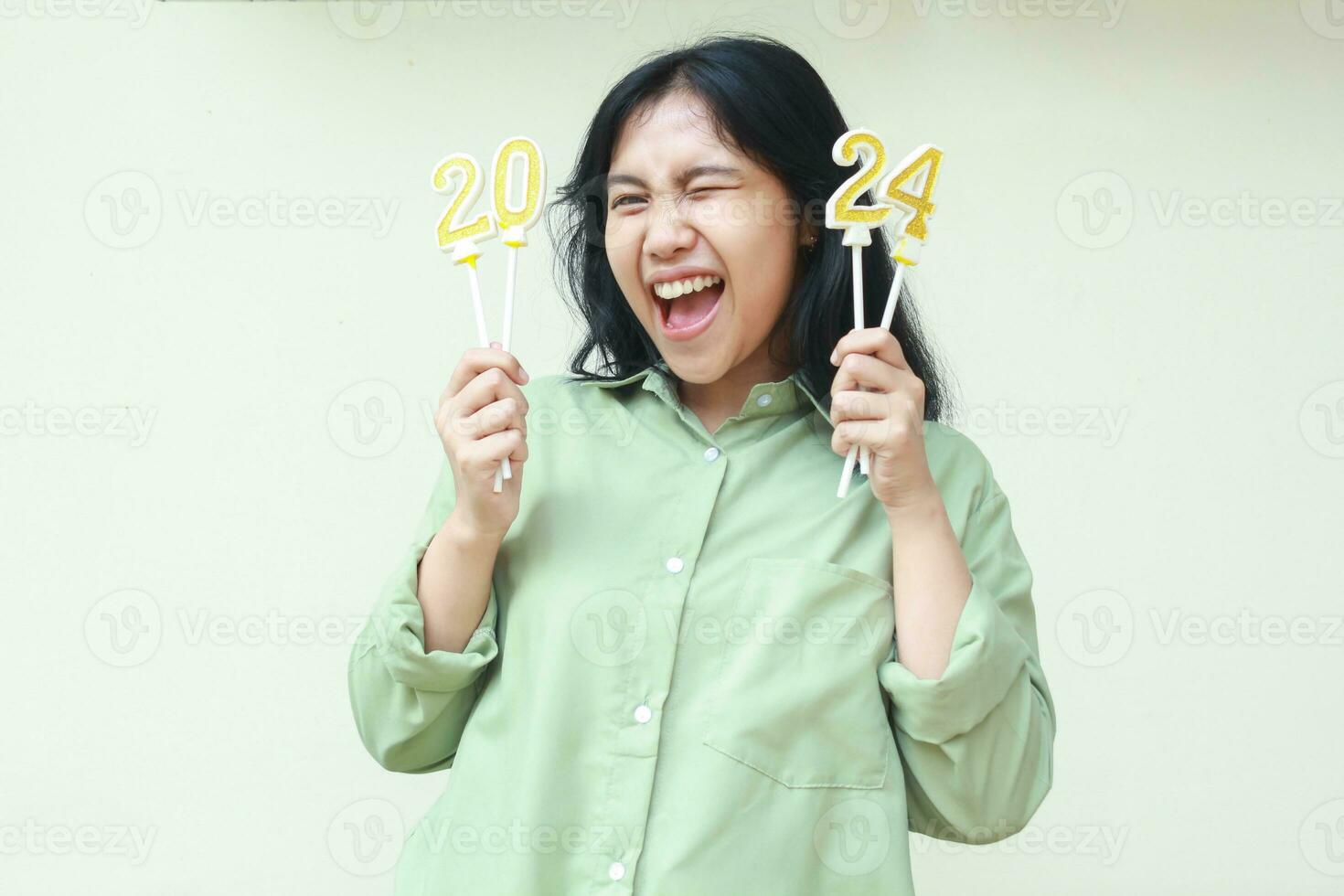 excited beautiful asian young woman screaming with yes gesture by raising hand holding gold numbers 2024 candles and blink an eye flirting to camera wear green shirt isolated. New years eve concept photo