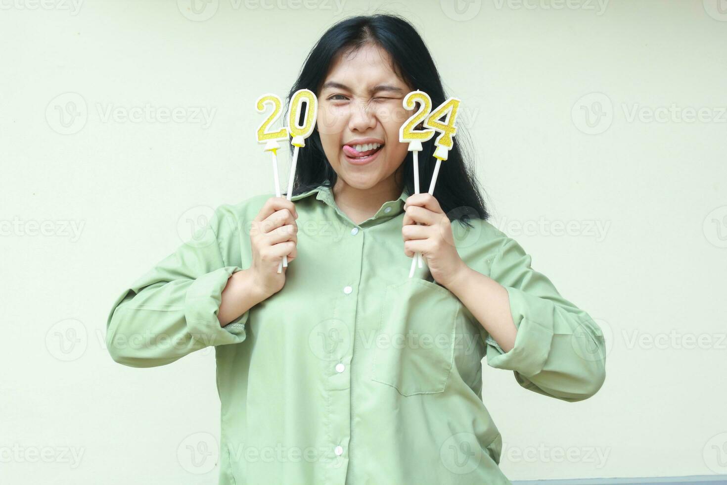 emocionado hermosa asiático joven mujer parpadeo ojos flirteador a cámara disfrutar 2024 nuevo años celebracion por participación oro reluciente número velas vistiendo verde terminado Talla camisa aislado en blanco antecedentes foto