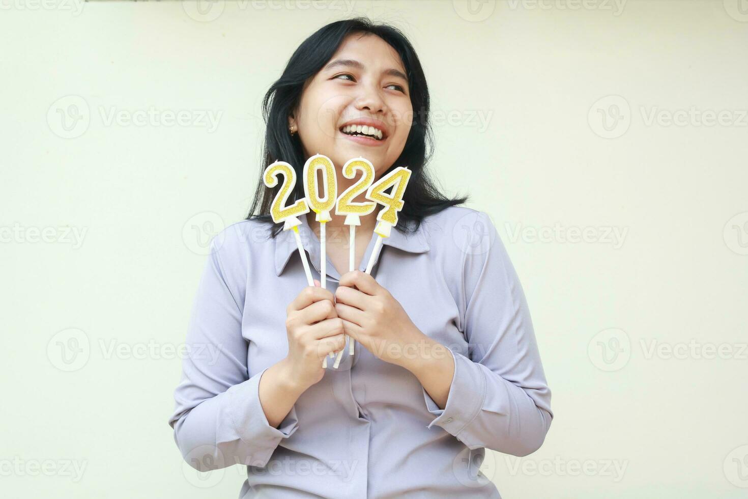 retrato de sonriente asiático joven negocio mujer emocionado para 2024 nuevo años celebracion Nueva York participación oro figura velas en mano vistiendo gris formal traje mirando lejos aislado en blanco antecedentes foto