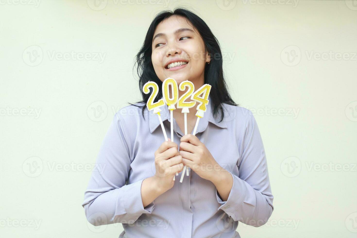 contento asiático joven mujer sonriente y deseando nuevo resolución en 2024 nuevo años víspera celebracion con participación número oro velas en mano vistiendo formal traje mirando lado aislado en blanco foto