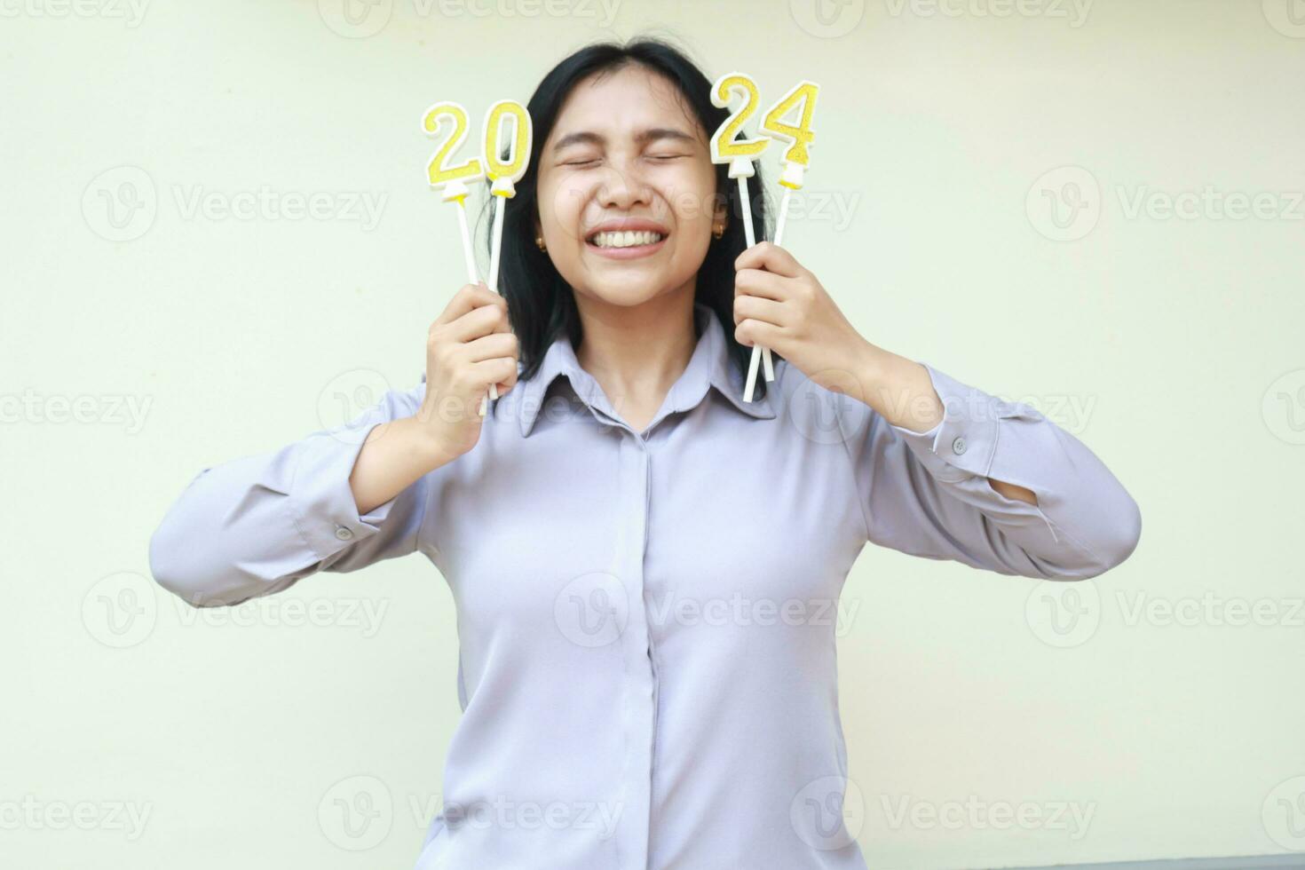 exitoso asiático joven negocio mujer emocionado en 2024 nuevo años víspera celebracion por participación dorado figura velas en manos y cerca ojos vistiendo gris formal camisa aislado por blanco antecedentes foto