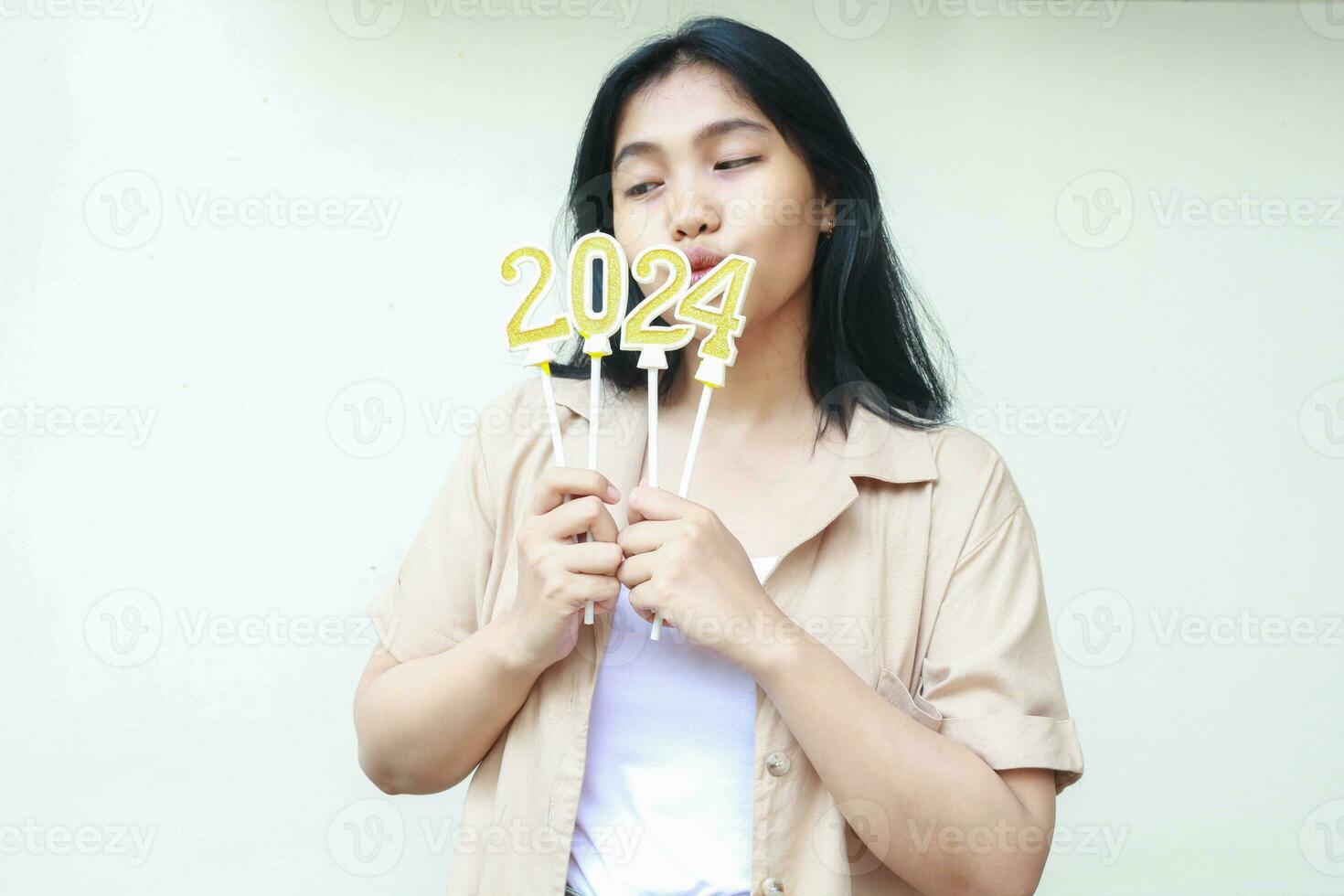 retrato de contento joven mujer asiático disfrutar 2024 nuevo años víspera celebracion mirando aparte a dorado dígito velas cubierta su boca, vestir casual marrón ropa aislado terminado blanco antecedentes foto