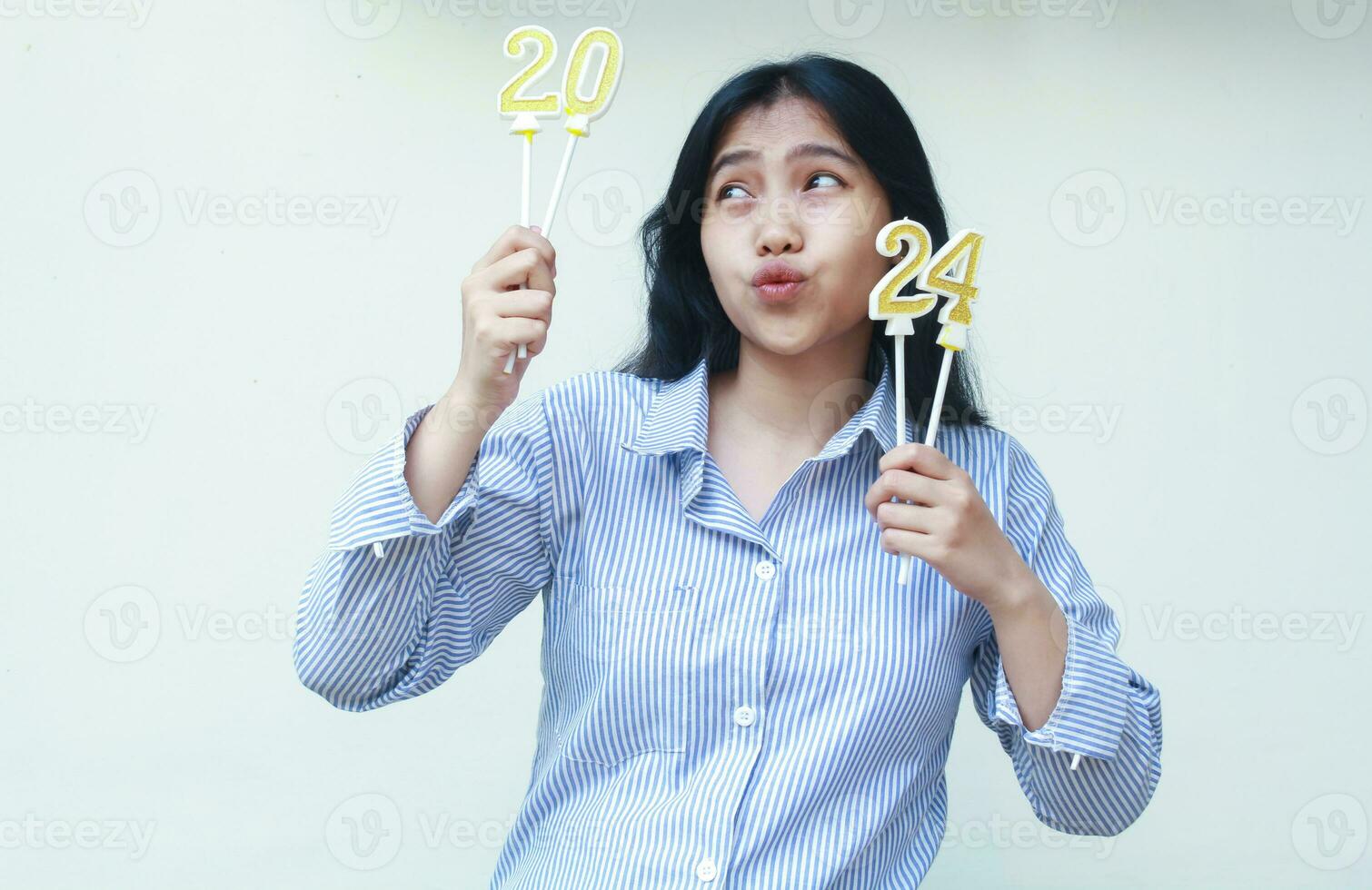 happy asian young business woman holding holding golden candles number 2024 and looking up with duck face wear blue stripes shirt isolated. beautiful female indonesian celebrate new years eve photo