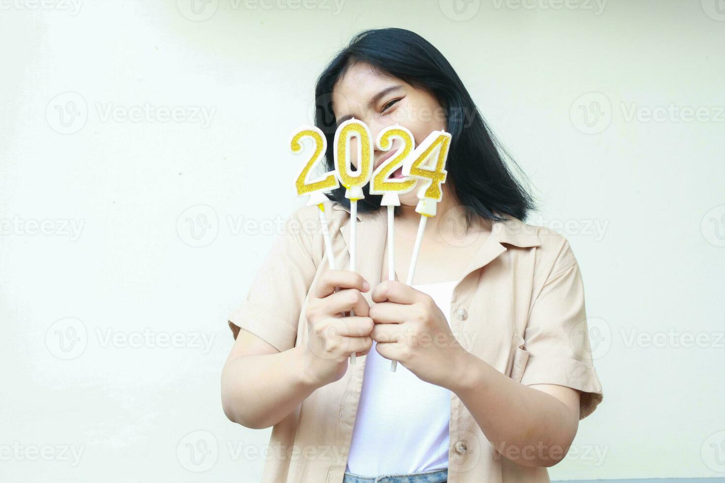 happy asian beautiful young woman smiling at camera to celebrate 2024 new years eve with golden number candles covering her face wear brown casual outer isolated on white background photo