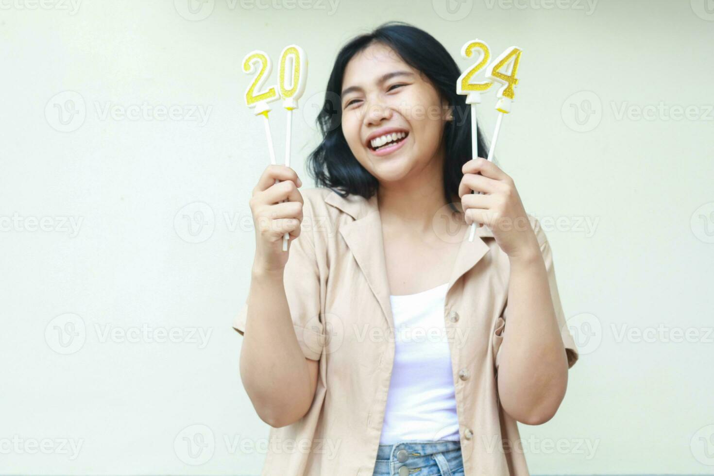 emocionado asiático joven mujer riendo contento y mirando lejos mientras sostener 2024 figura velas vestir de moda marrón exterior y mezclilla pantalones aislado en blanco foto