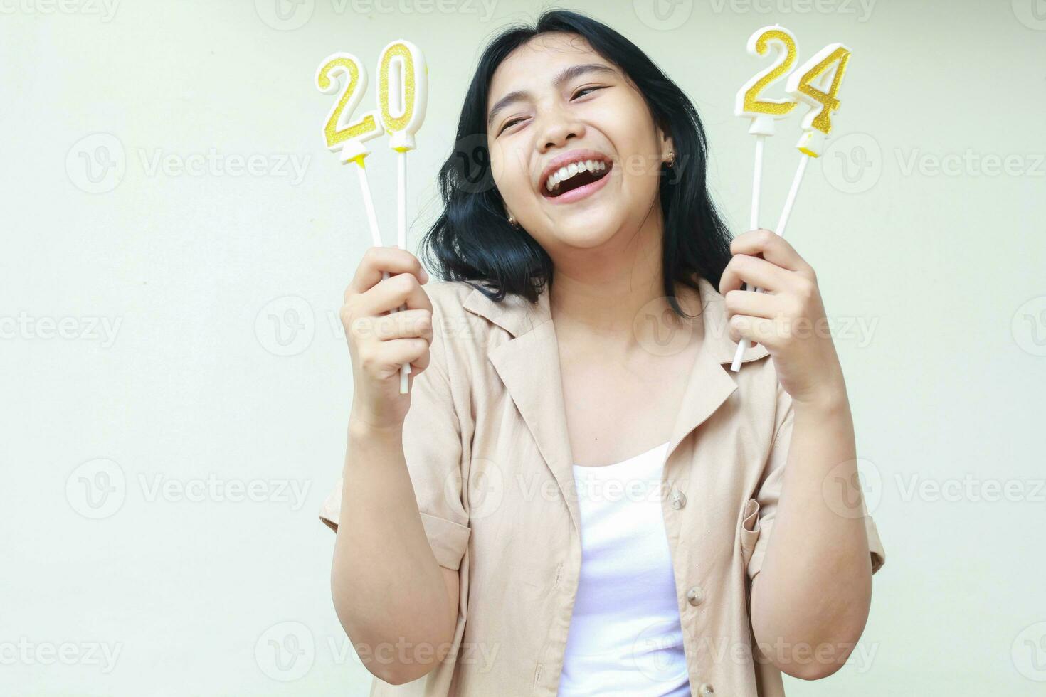 retrato de despreocupado hermosa asiático joven mujer riendo mientras sostener reluciente 2024 velas con con dientes sonrisa y mirando aparte vestir de moda casual marrón exterior aislado en blanco foto