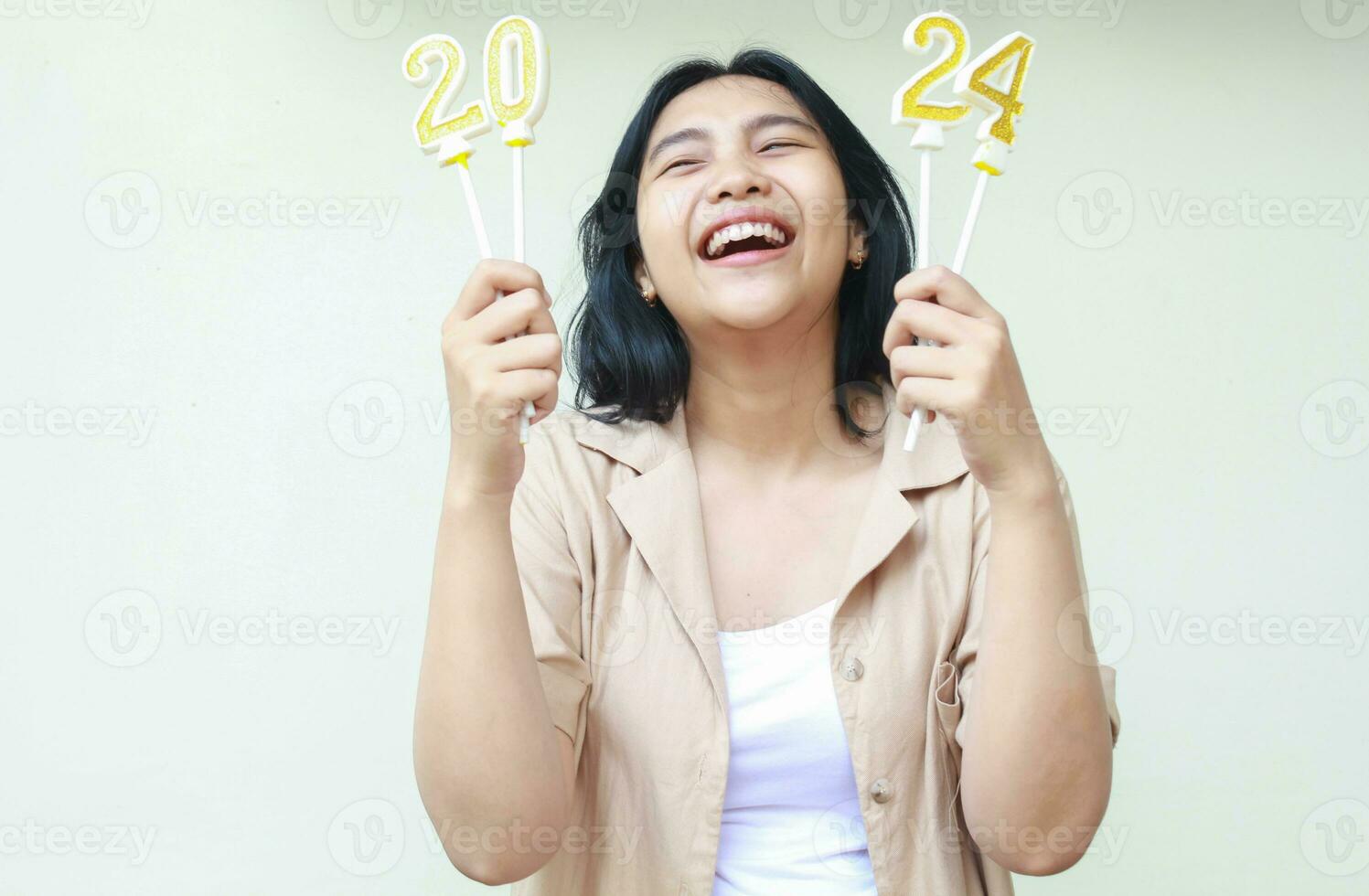 alegre asiático joven mujer riendo disfrutar 2024 nuevo años víspera fiesta con participación número velas vistiendo casual marrón exterior, mirando arriba, aislado terminado blanco antecedentes foto