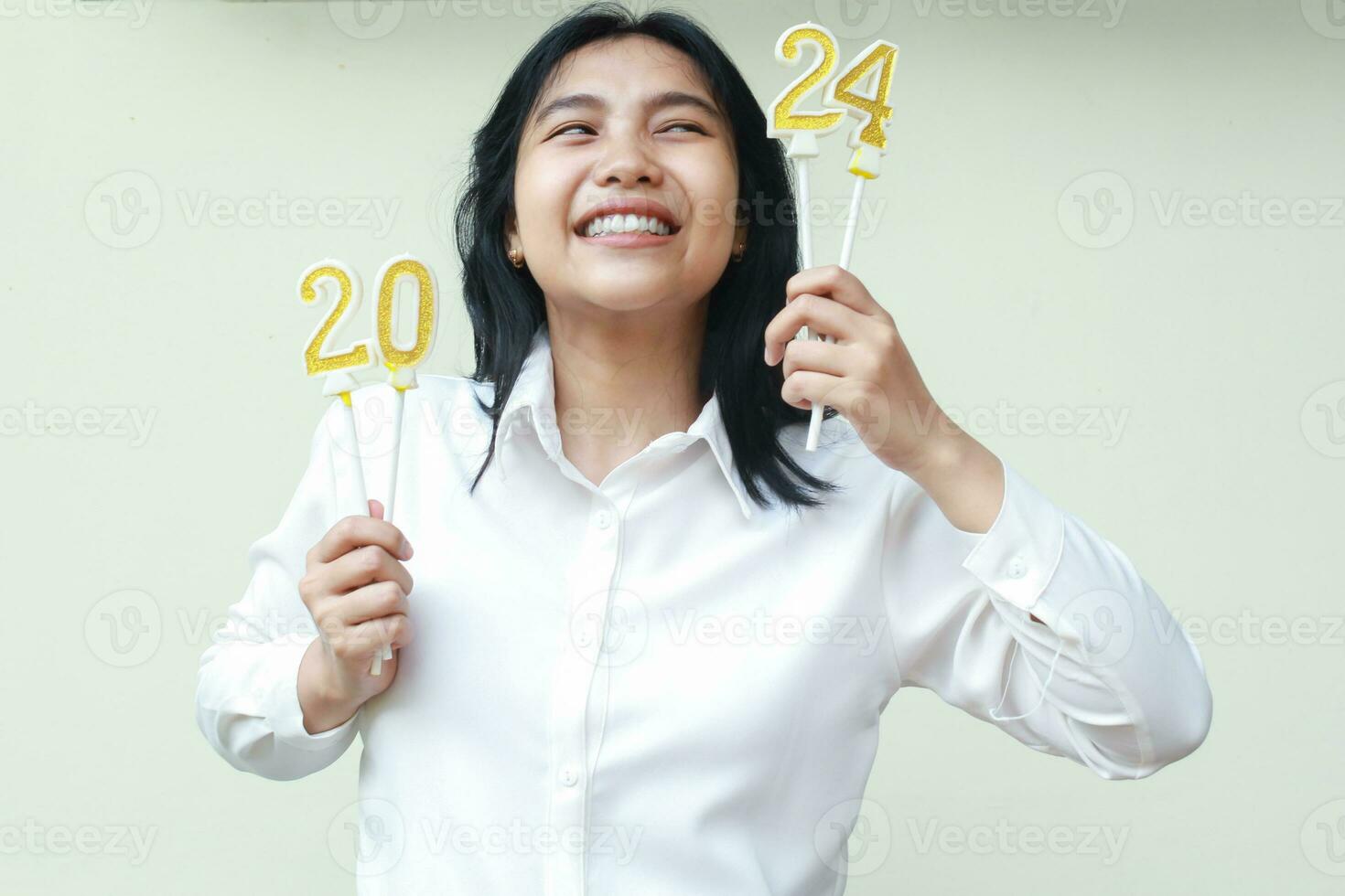 emocionado asiático joven mujer compañero de trabajo sonriente y mirando aparte a número 24 de 2024 figura velas sostener en levantado mano vestir blanco camisa, aislado, hembra deseo nuevo resolución en nuevo años víspera foto