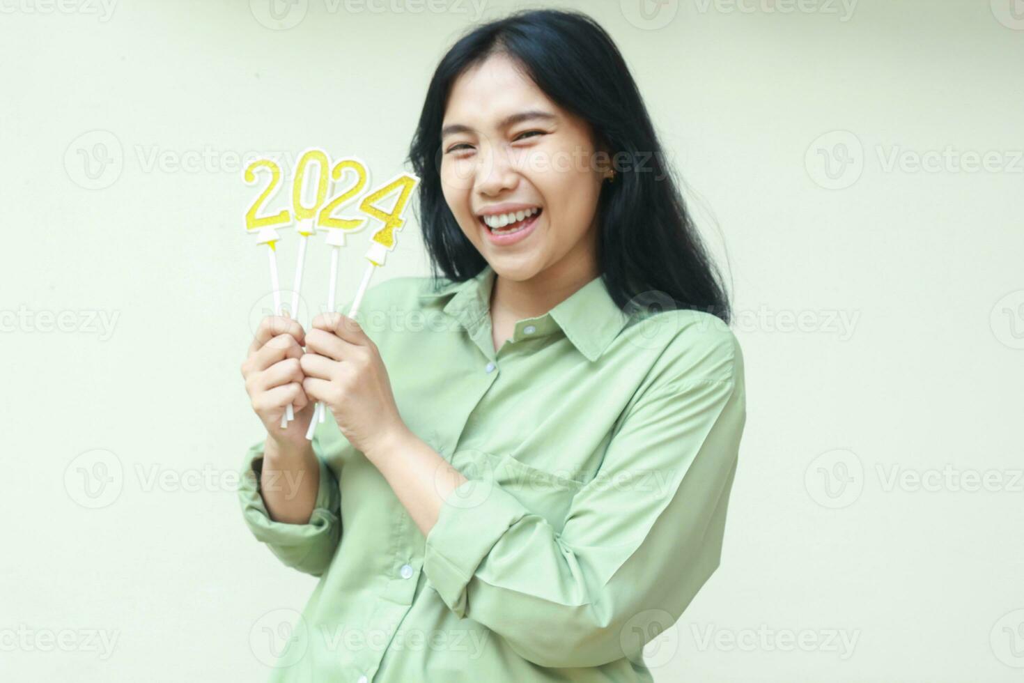 alegre asiático joven mujer sonriente a cámara vistiendo verde casual ropa con levantamiento brazo participación 2024 figura velas a celebrar nuevo años víspera, aislado terminado blanco antecedentes foto