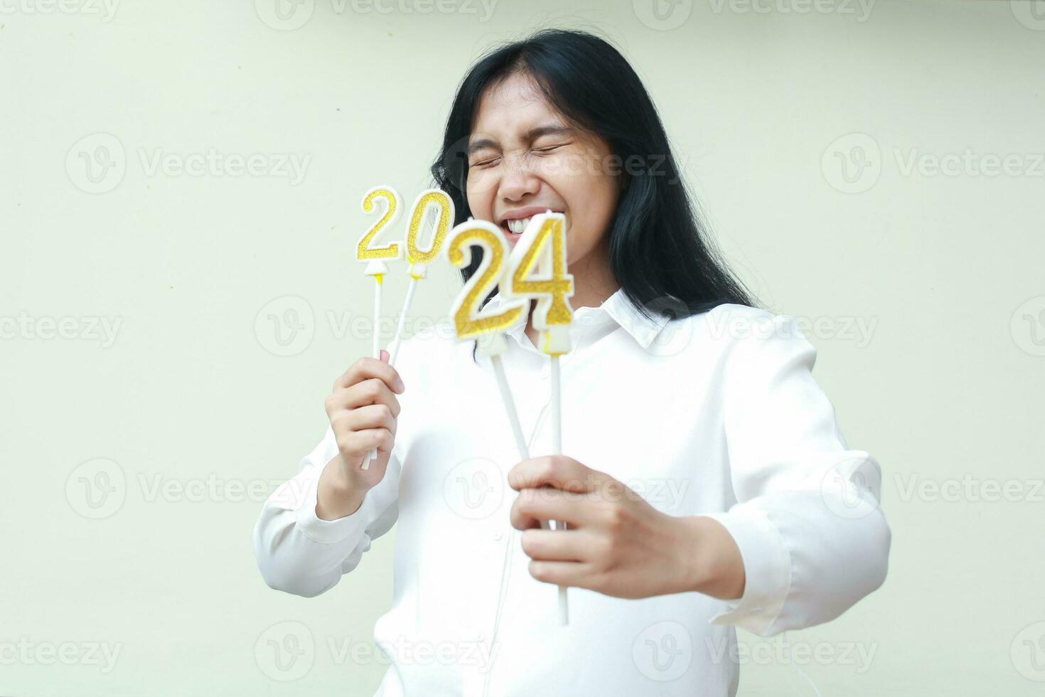 excited asian young woman coworker smiling with closed eye and showing 2024 figure candles to camera wearing formal white shirt, isolated, new years eve concept photo