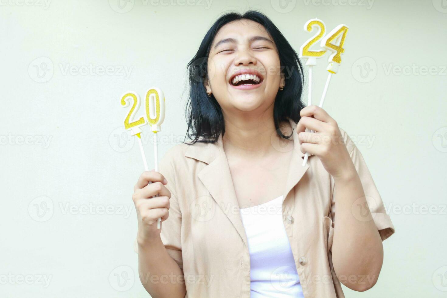 riendo hermosa asiático joven mujer participación dorado velas número 2024 en manos con cerrado ojos y con dientes sonrisa disfrutar celebrando nuevo años víspera vestir elegante casual marrón exterior aislado en blanco foto