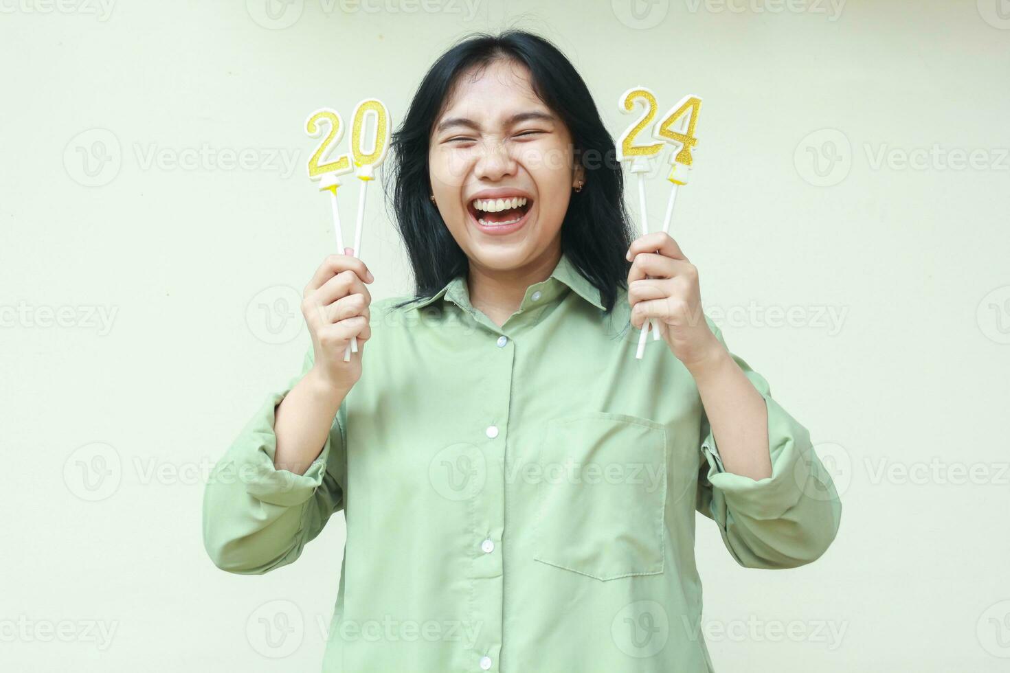 retrato de emocionado asiático joven mujer hipster riendo con cerrado ojo y levantando brazo con 2024 figura velas sostener en mano vistiendo verde terminado Talla camisa, aislado en blanco. nuevo años víspera concepto foto