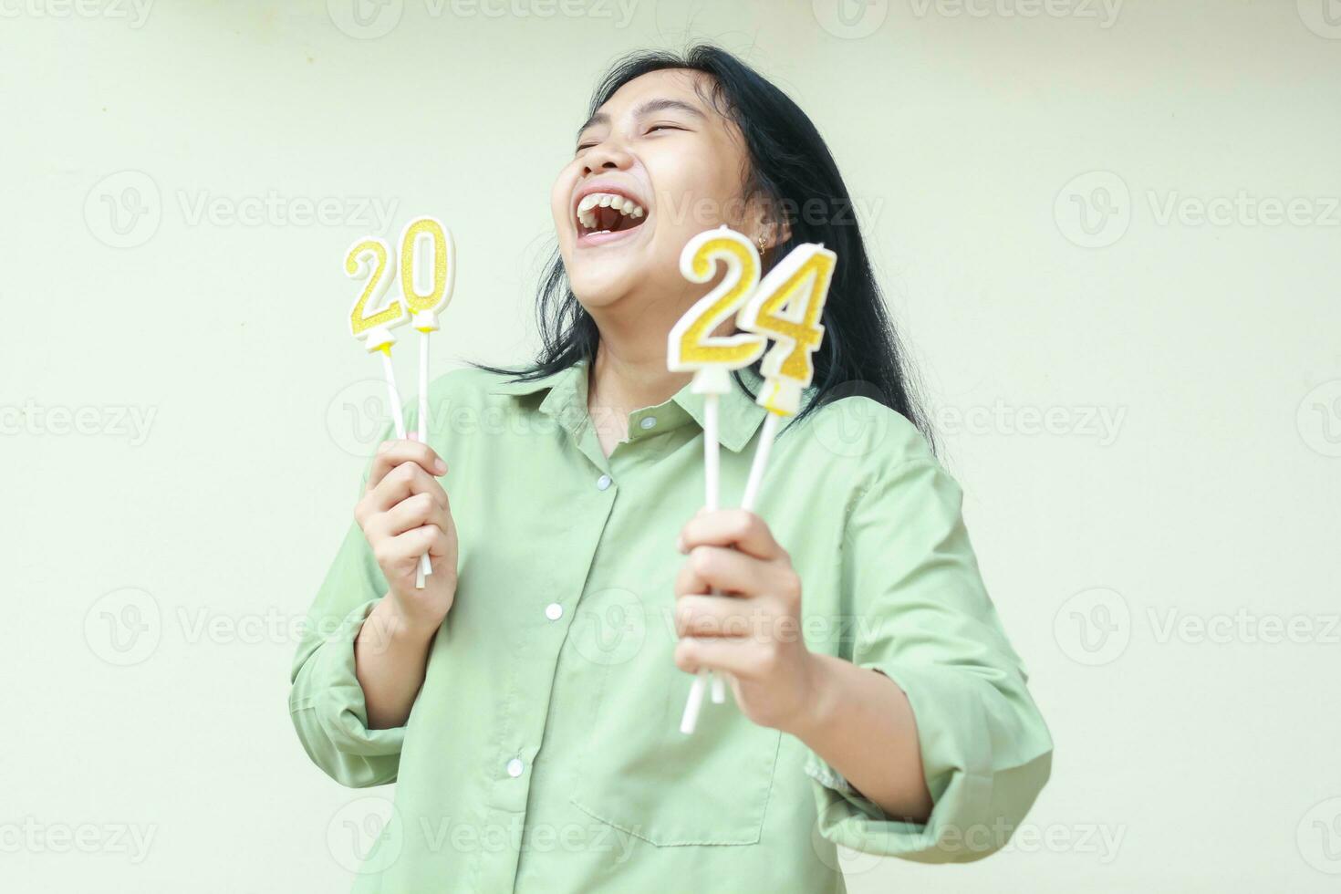 carefree asian young woman laughing and looking away wearing green oversize shirt with raising arm showing 2024 figure candles to camera, isolated, female celebrate new years eve photo