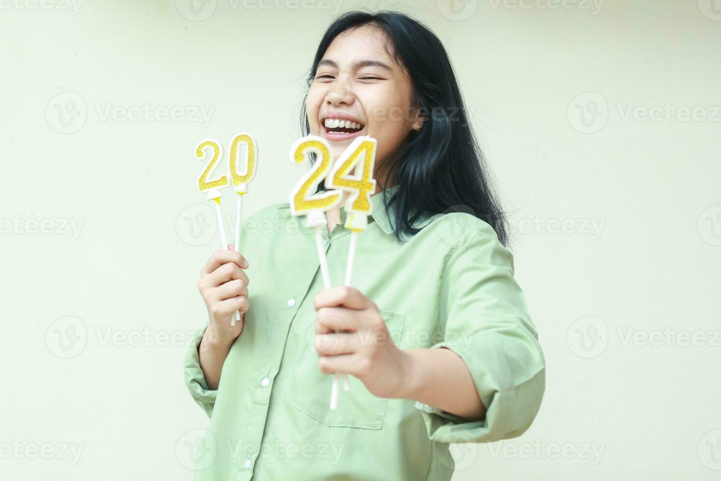 retrato de despreocupado asiático joven mujer riendo a cámara con cerrado ojo y espectáculo 2024 figura velas levantamiento en mano vistiendo verde terminado Talla Shir aislado en blanco antecedentes. mirando a cámara foto