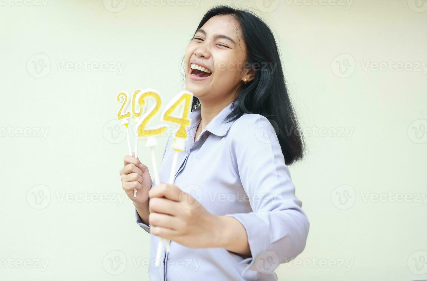 portrait of excited asian young woman coworker laughing at camera with closed eye and show 2024 figure candles lifting on hand wear grey formal shirt, isolated on white, new years eve concept photo