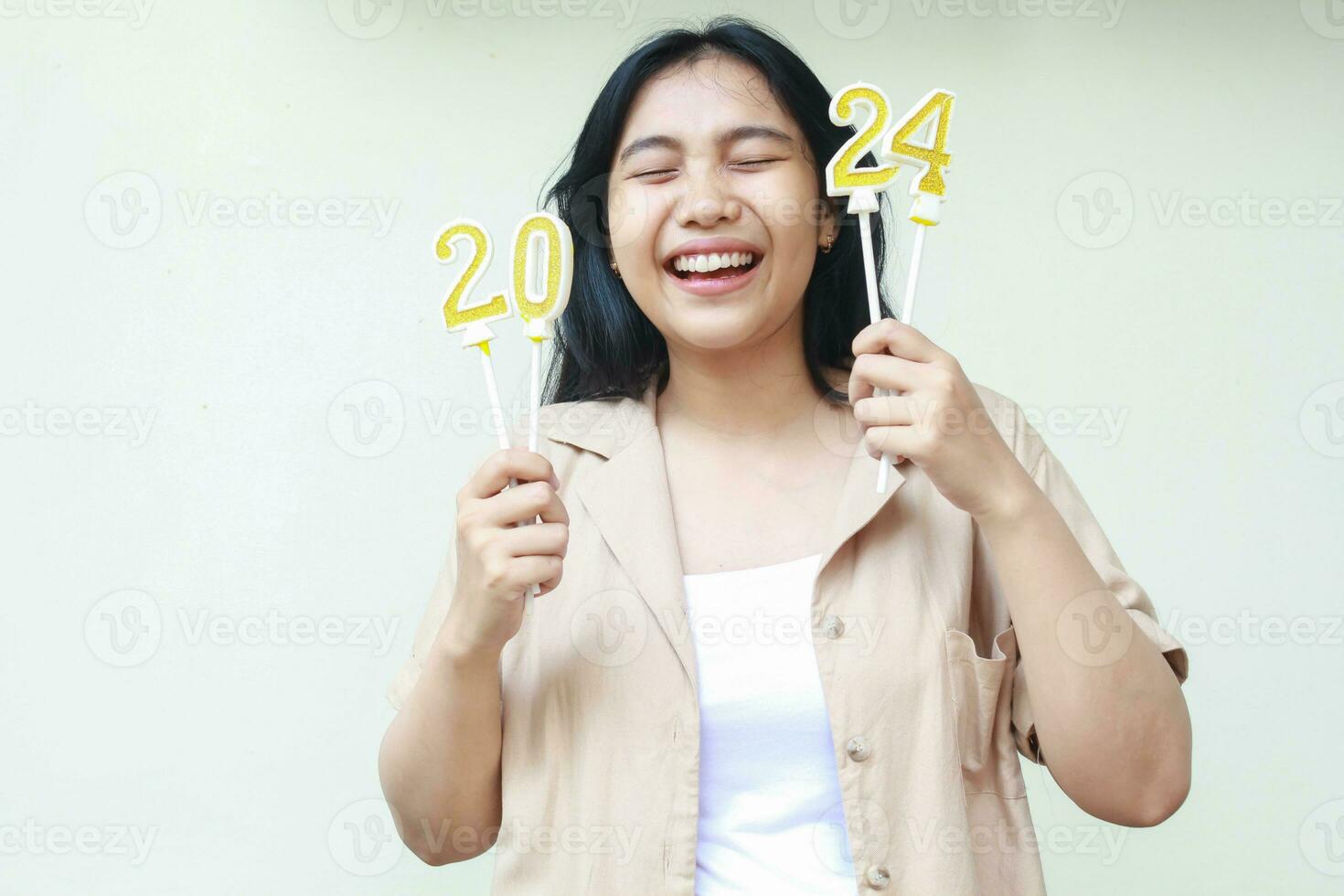 atractivo asiático joven mujer riendo con ojo cerrado y levantamiento brazo participación 2024 dorado velas vistiendo casual marrón exterior celebrando nuevo años víspera aislado en blanco antecedentes foto