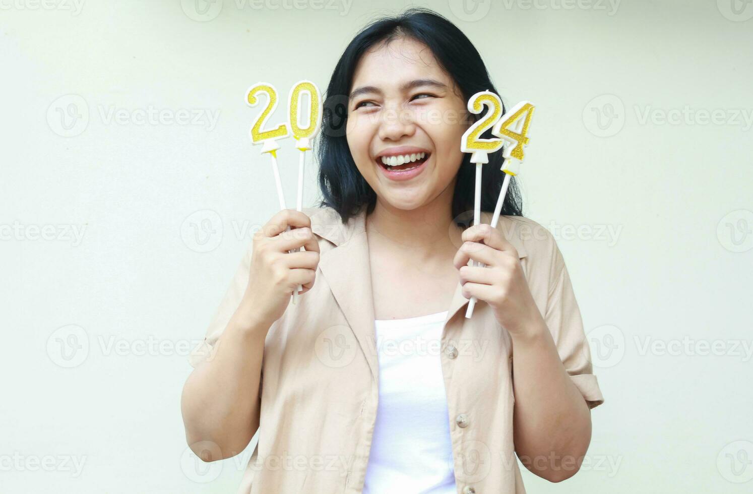 riendo asiático joven mujer participación 2024 oro velas y mirando aparte con con dientes sonrisa vistiendo marrón casual exterior aislado en blanco antecedentes foto