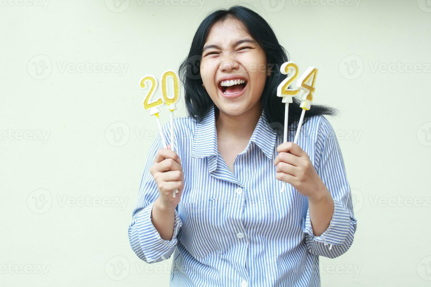 portrait of excited digital nomad laughing at camera with closed eye and open mouth while holding 2024 figure candles wear blue stripes shirt isolated on white. new year eve celebration concept photo