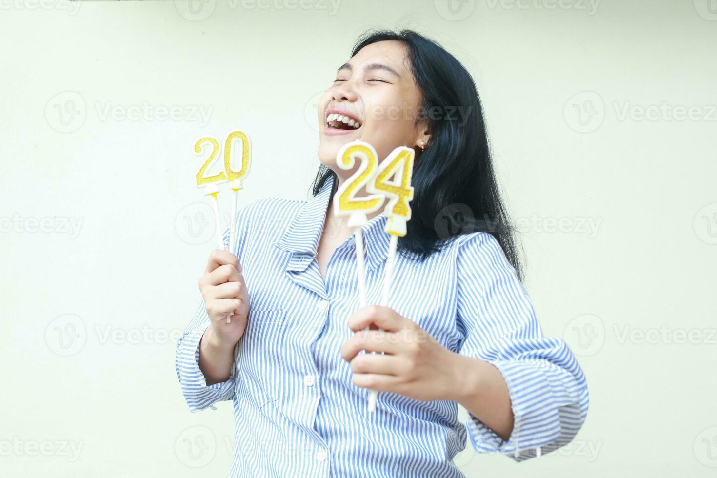 cheerful asian young woman celebrating new years eve with laugh and happy smile, looking up with closed eye, female lifting arm hold 2024 figure candles wear blue stripes fashionable clothes isolated photo