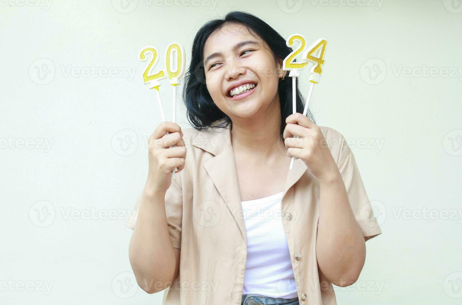 cheerful young woman of asian ethnicity laughing and looking aside with bright smile with raising arm holding 2024 figure candles wear stylish brown outer isolated on white photo