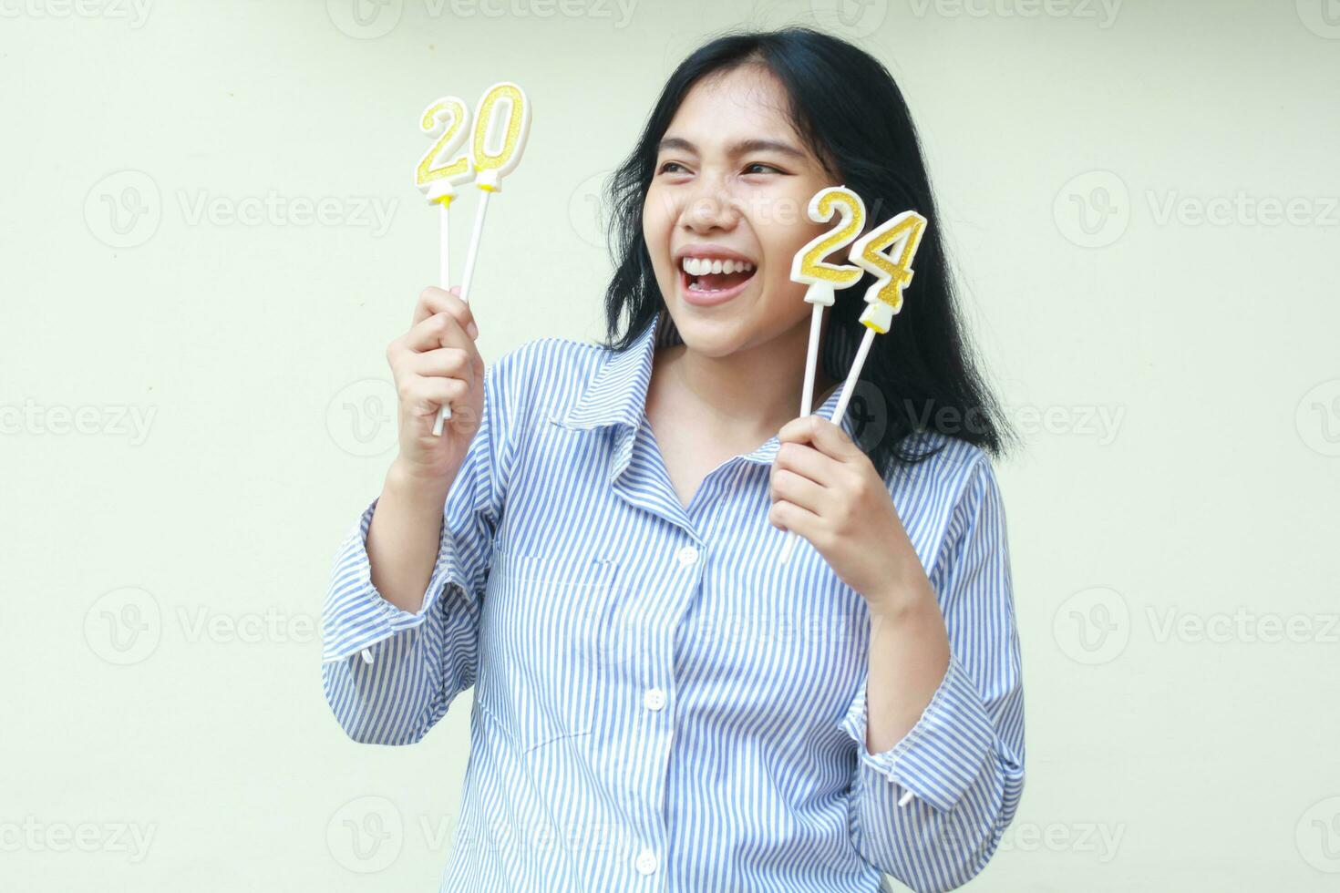 playful young woman asian remote worker looking away with toothy smile while raising arm hold 2024 candles on hand wear blue stripes clothes isolated on white photo