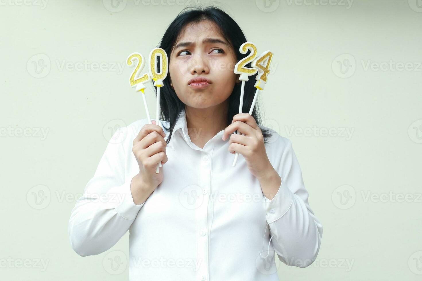 thoughtful asian young woman holding 2024 figure candle with curiosity and looking aside to number 24 wearing white formal shirt, isolated photo